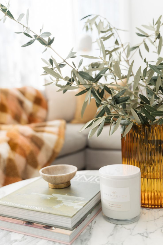 fall-inspired coffee table decor with amber glass vase filled with olive branches, stacked coffee table books, neutral ceramic bowl, and white candle in a cozy living room

