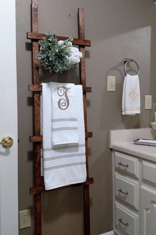 Bathroom featuring a dark wood towel ladder leaning against a taupe wall, styled with monogrammed white towels, faux greenery in a wire basket, and complemented by a nearby vanity with classic white cabinetry and chrome hardware. 