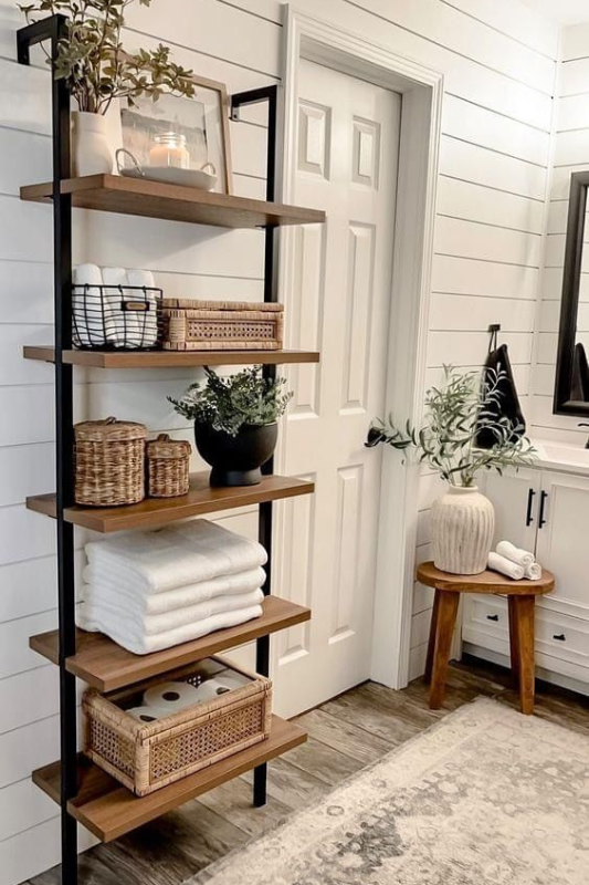Bathroom styled with a modern farmhouse open shelving unit featuring wood and black metal, organized with white towels, woven baskets, greenery, and candles, set against a white shiplap wall and rustic wood flooring. 