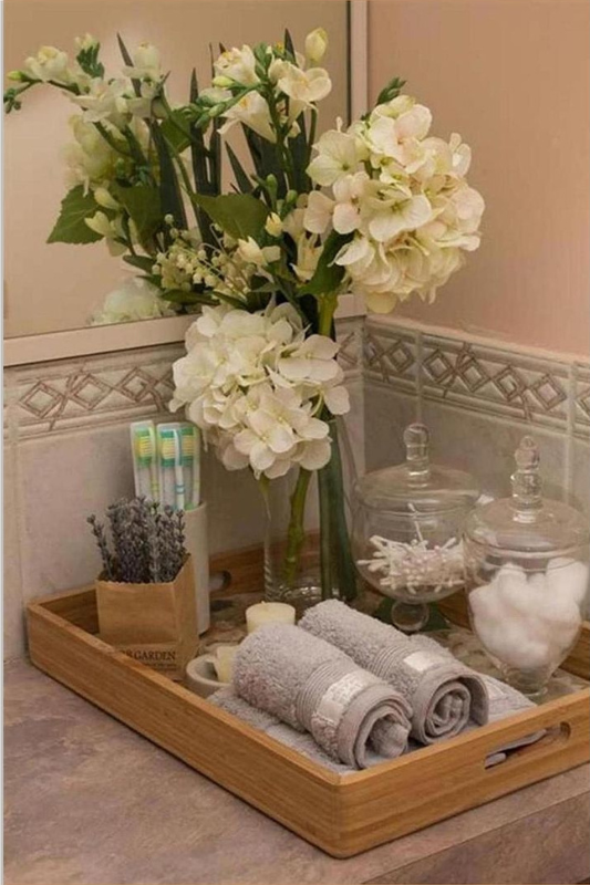 Elegant bathroom vanity tray decor featuring rolled gray towels, a tall glass vase with white hydrangeas, apothecary jars with cotton balls and swabs, lavender stems, and toothbrushes, all styled on a wooden tray for a spa-inspired look. 