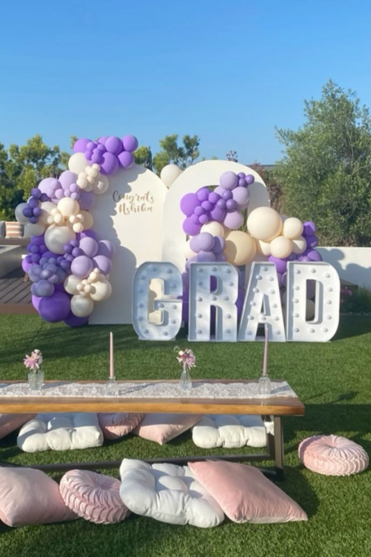 Outdoor graduation party setup with pastel purple and cream balloon backdrop, large GRAD marquee letters, and a low picnic table surrounded by soft pink and white cushions. 