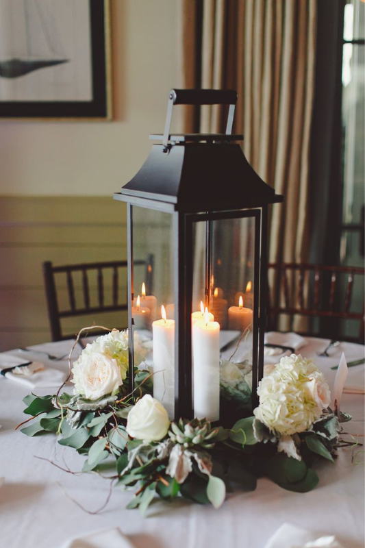 Elegant black lantern centerpiece featuring glowing white pillar candles surrounded by a lush floral ring with white roses, hydrangeas, succulents, and silver dollar eucalyptus on a round dining table with a white tablecloth. 