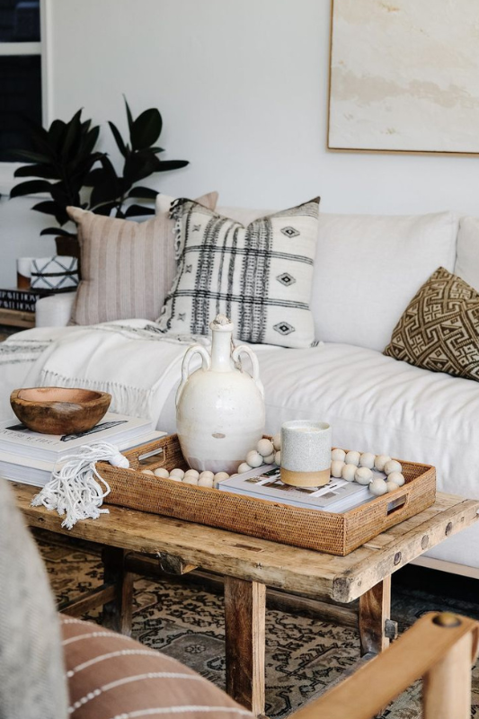 boho-inspired coffee table styling with a woven basket tray, ceramic jug, wooden bead garland, neutral books, and cozy earth-toned pillows in a relaxed living room setting