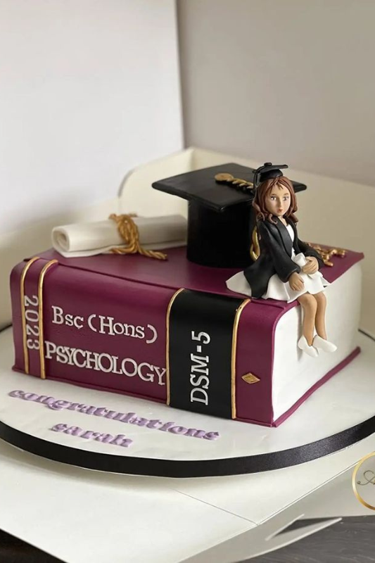 Graduation cake designed as a psychology textbook with a fondant figure of a graduate, a diploma scroll, and a graduation cap
