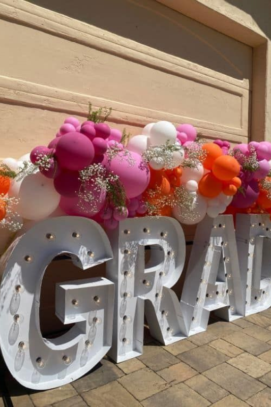 Outdoor graduation party decor featuring large marquee GRAD letters with lightbulbs, decorated with pink, orange, and white balloons and baby's breath florals for a vibrant 2025 celebration.