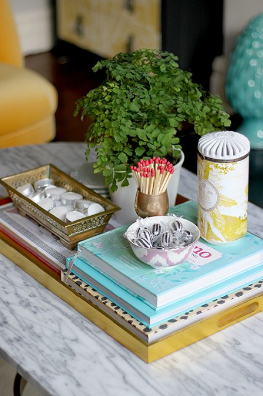vibrant coffee table styling with stacked colorful books, a mini potted plant, a matchstick holder, tea light candles, and a decorative spa tin on a marble table

