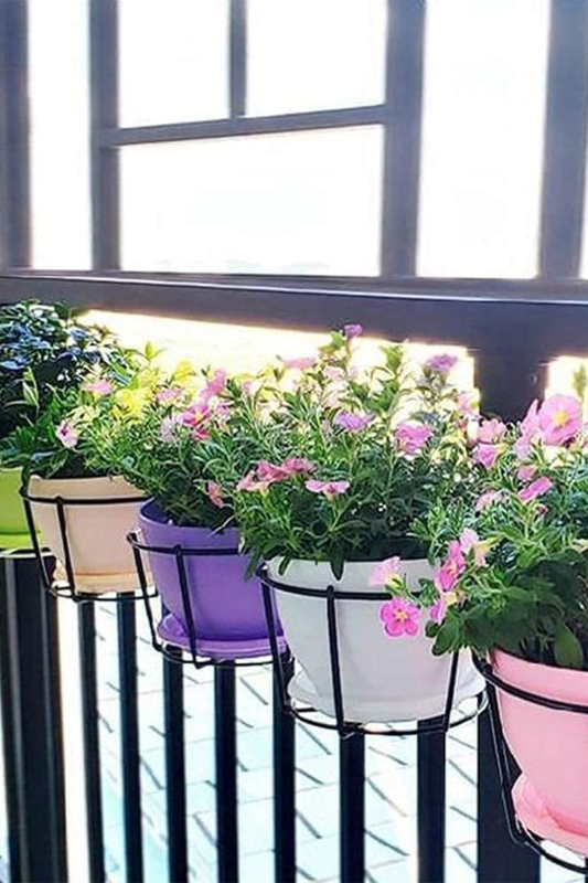Row of colorful hanging flower pots in pastel shades—pink, purple, white, and green—mounted on a black balcony railing, filled with blooming pink flowers and greenery. 