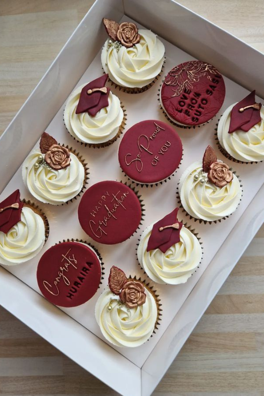 Box of elegant graduation cupcakes with ivory frosting, maroon fondant toppers featuring caps, floral accents, and congratulatory messages like “So Proud of You” and “Happy Graduation.”