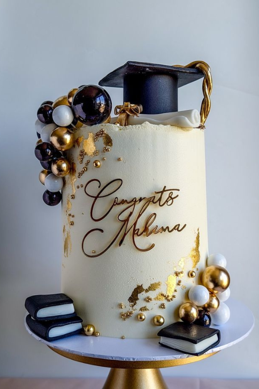 Tall white graduation cake with gold accents, topped with a black graduation cap, decorative spheres, edible books, and the inscription
