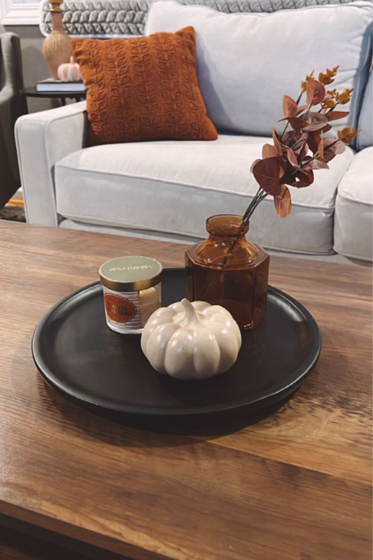 cozy fall coffee table decor with a white ceramic pumpkin, pumpkin bread candle, and amber glass vase on a black tray in a warm living room

