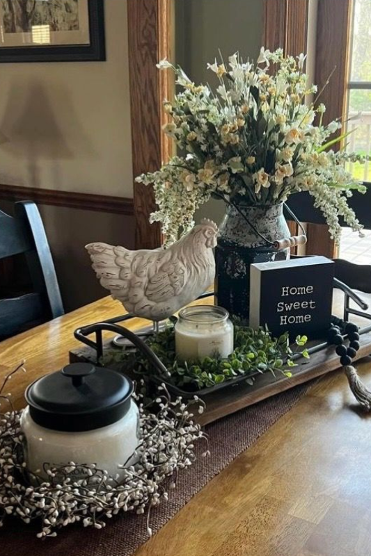 Farmhouse dining table centerpiece featuring a ceramic chicken figurine, a floral arrangement in a vintage-style pitcher, a “Home Sweet Home” sign, candles, greenery, and wooden bead garland, all styled on a rustic wooden tray with a cozy cottage charm. 