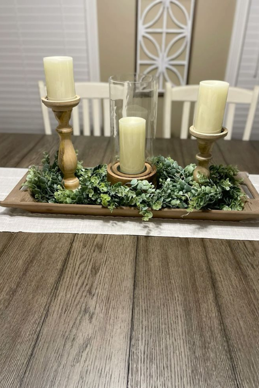 Rustic dining table centerpiece featuring three cream pillar candles on wooden holders, surrounded by lush faux greenery on a rectangular wooden tray, styled on a dark wood table with a neutral table runner. 
