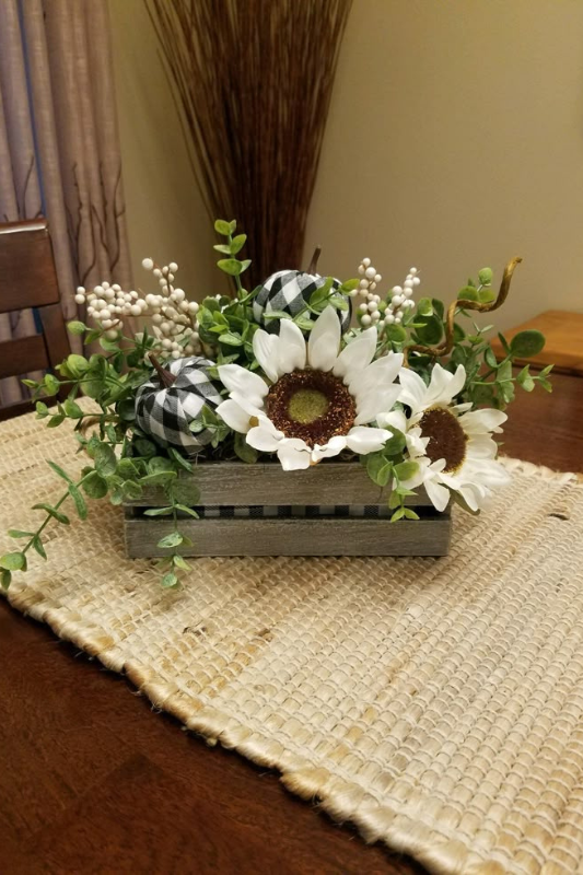 Rustic farmhouse centerpiece featuring white sunflowers, greenery, faux white berries, and black-and-white buffalo check pumpkins arranged in a gray wooden crate, styled on a natural woven table runner. 