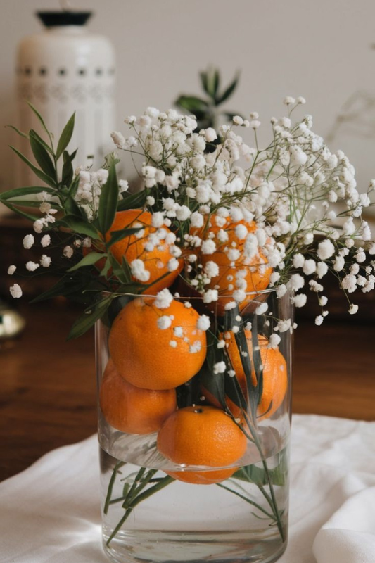 Clear glass vase filled with fresh oranges, delicate baby’s breath flowers, and greenery, styled as a vibrant and refreshing table centerpiece on a white cloth runner. 
