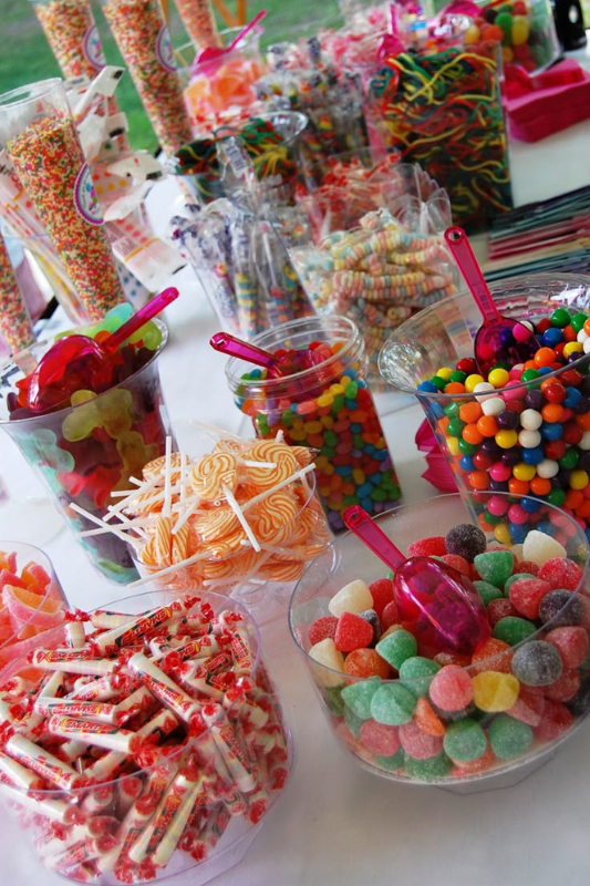 A colorful graduation candy table with jars filled with assorted sweets including lollipops, gummy bears, Smarties, jelly beans, and rainbow candies, styled with scoops for a self-serve dessert station.