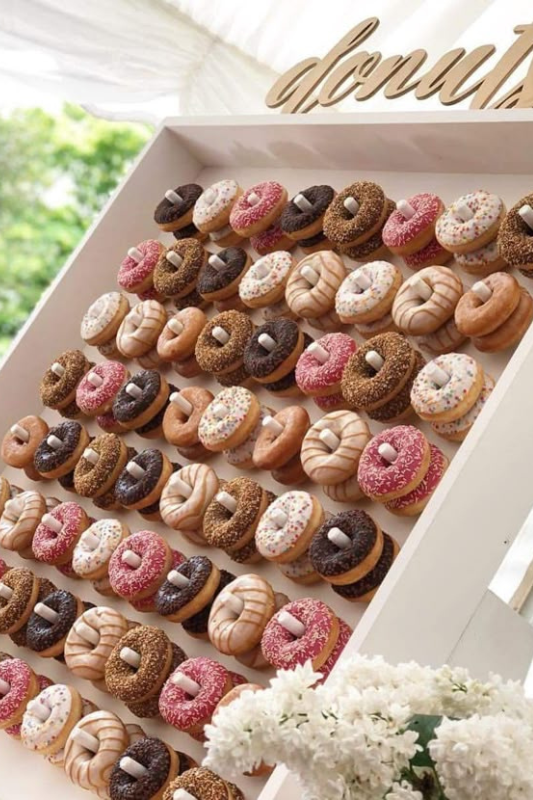 Graduation party donut wall featuring rows of assorted donuts with sprinkles, glaze, and chocolate toppings, displayed on a white board with a wooden “donuts” sign at the top.