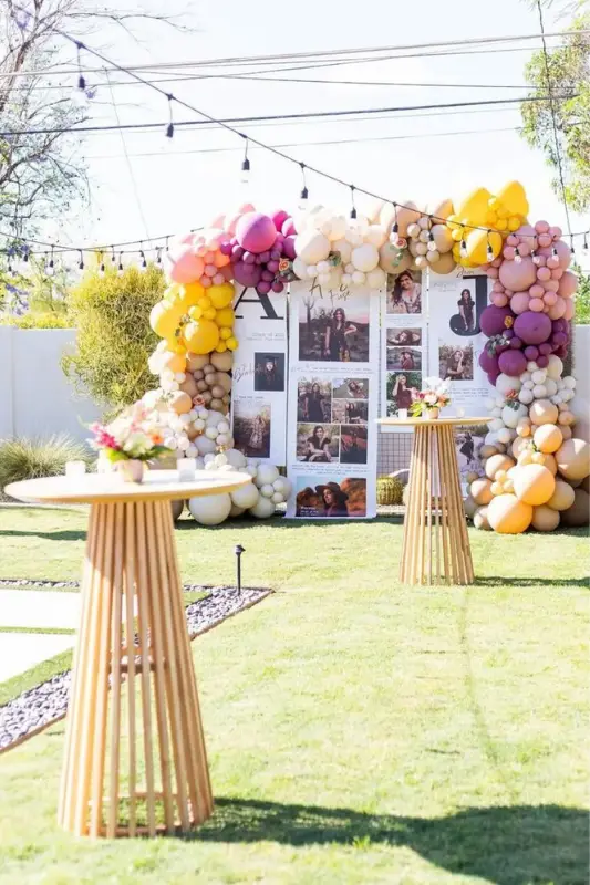 Outdoor graduation party photo collage backdrop decorated with colorful pastel balloons in purple, pink, yellow, and cream, surrounded by high-top tables and floral arrangements.