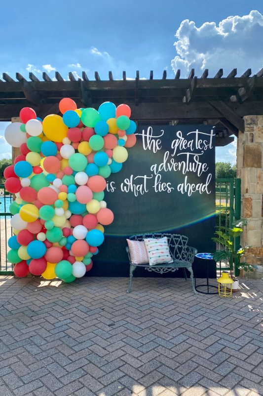 Colorful graduation photo booth backdrop featuring a vibrant balloon arch and motivational quote 'The greatest adventure is what lies ahead,' styled with modern outdoor seating.