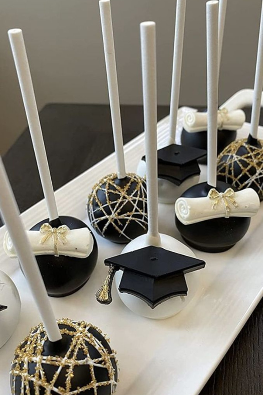 Black and white graduation cake pops decorated with gold bows, diploma scrolls, and fondant graduation caps, arranged on a white rectangular tray.