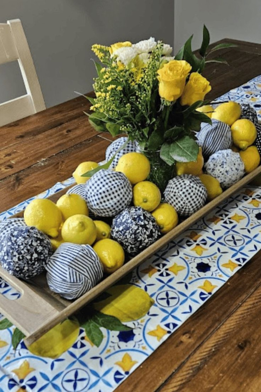 Vibrant dining table centerpiece featuring a wooden tray filled with fresh lemons, blue-and-white patterned fabric balls, and a clear vase of yellow and white flowers, styled on a bold patterned table runner for a cheerful spring or summer look. 