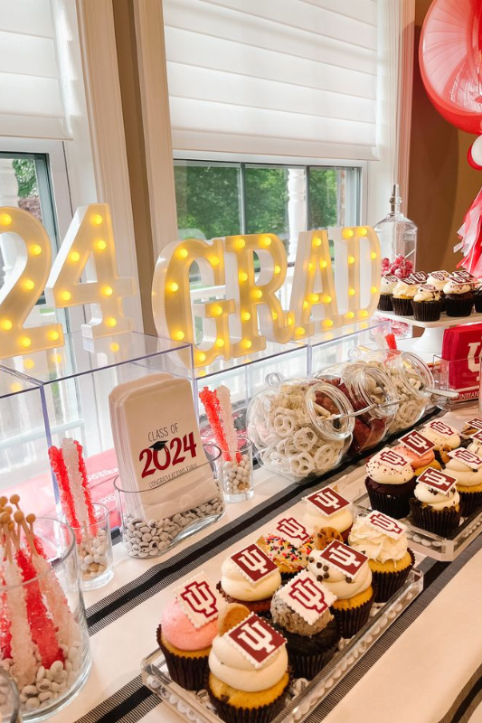 A letter-shaped graduation cake decorated with blue and gray frosting, chocolate pieces, strawberries, a diploma topper, and a miniature graduation cap, displayed outdoors on a white board.