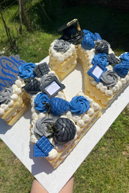 A letter-shaped graduation cake decorated with blue and gray frosting, chocolate pieces, strawberries, a diploma topper, and a miniature graduation cap, displayed outdoors on a white board.
