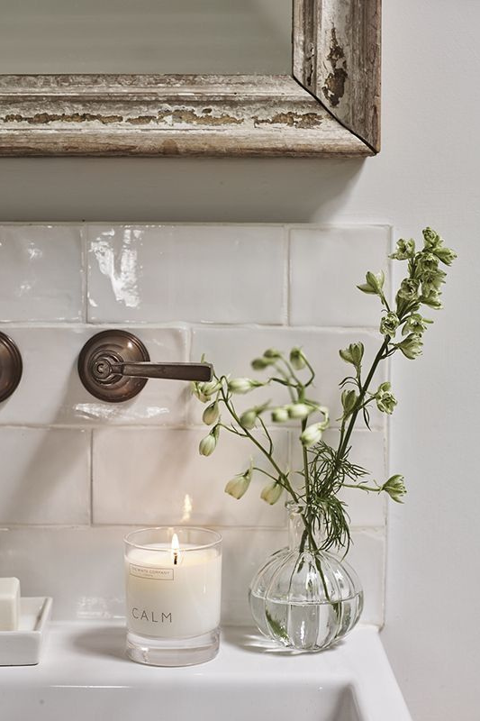 Minimalist bathroom sink area featuring a vintage wood-framed mirror, white subway tile backsplash, a lit "Calm" candle, a clear glass vase with fresh greenery, and brushed bronze wall-mounted faucets for a serene, rustic touch. 