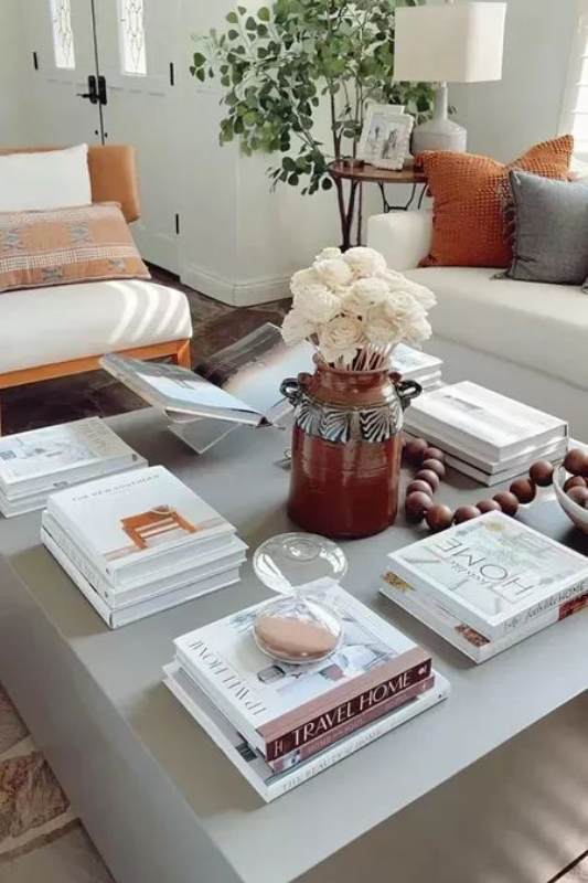 stylish modern coffee table with stacked home decor books, a terracotta vase of white flowers, wooden beads, and cozy throw pillows in a bright neutral living room

