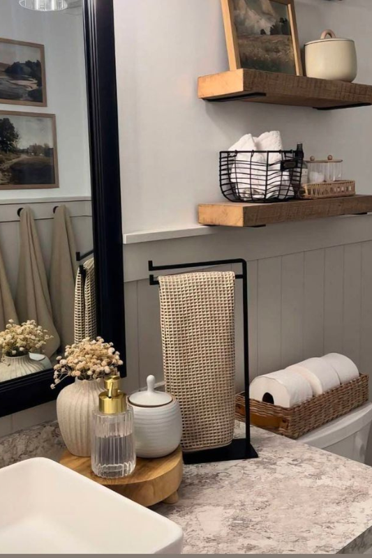 Modern farmhouse bathroom featuring open wood shelves with baskets and artwork, a black-framed mirror, textured hand towel on a metal stand, and natural decor accents like ceramic containers and dried florals on a marble-look vanity. 