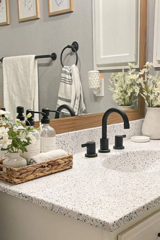 Modern neutral bathroom featuring a speckled countertop, matte black faucet and hardware, wood-framed mirror, and decorative touches like a woven tray with soap and flowers, white ceramic vase, and clean striped towels for a fresh, styled look. 