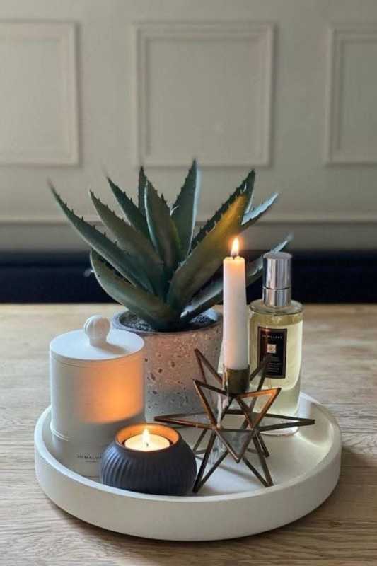 Contemporary round tray centerpiece featuring a faux succulent in a textured planter, two decorative candles, a geometric candle holder with a taper candle, and a luxury room spray, all arranged on a natural wood surface for a cozy, modern aesthetic. 