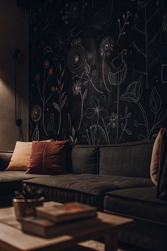 Cozy modern living room with a dark sectional sofa, blush and rust throw pillows, and a dramatic black accent wall featuring hand-drawn floral artwork in soft lighting 