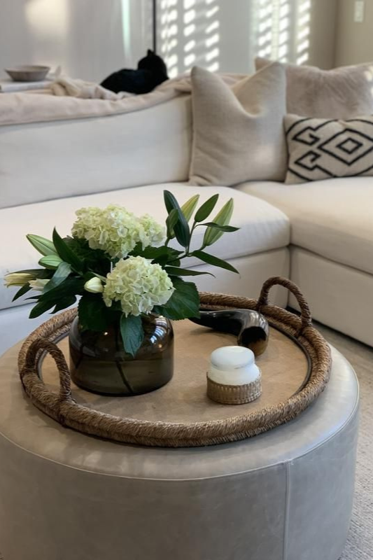 cozy living room ottoman styled with a woven tray, white hydrangea arrangement in a glass vase, and a candle for soft neutral decor

