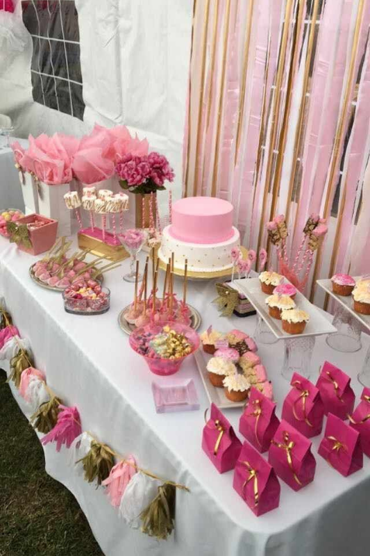 Pink and gold-themed graduation dessert table featuring a pink cake, cupcakes, dipped pretzels, marshmallow pops, favor bags, and floral decorations with a shimmering pink and gold backdrop.