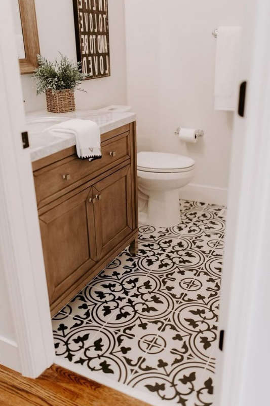 Stylish powder room featuring bold black and white patterned floor tiles, a warm wood vanity with marble countertop, a white toilet, and minimal decor including a potted plant and framed typography wall art. 