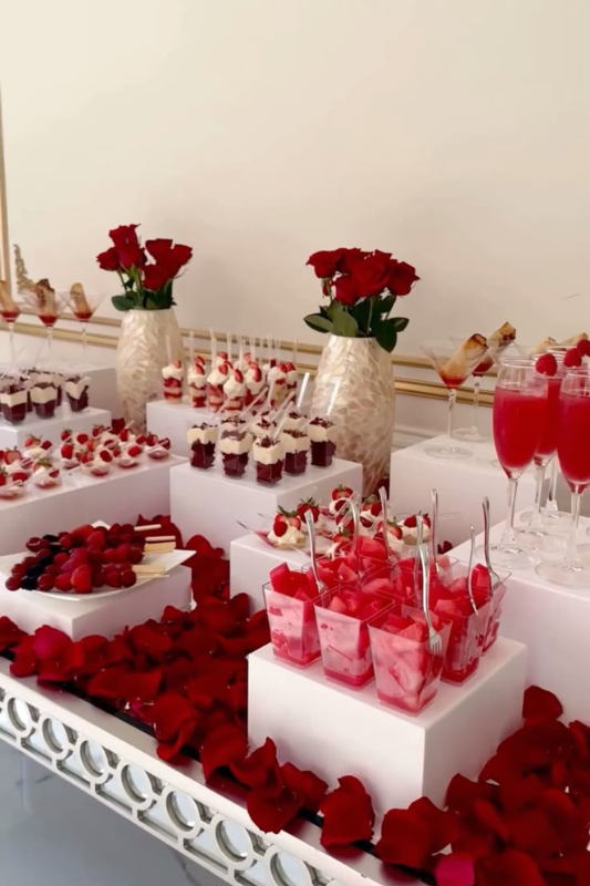 Elegant red and white graduation dessert table with assorted mini desserts, berry skewers, mousse cups, and champagne glasses, decorated with red rose petals and floral centerpieces.