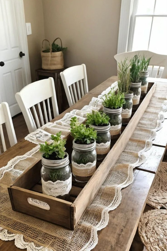 Farmhouse-style dining table centerpiece featuring mason jars wrapped in burlap and lace, each filled with fresh herbs and greenery, arranged inside a long wooden tray atop a burlap and lace table runner on a wooden farmhouse table. 