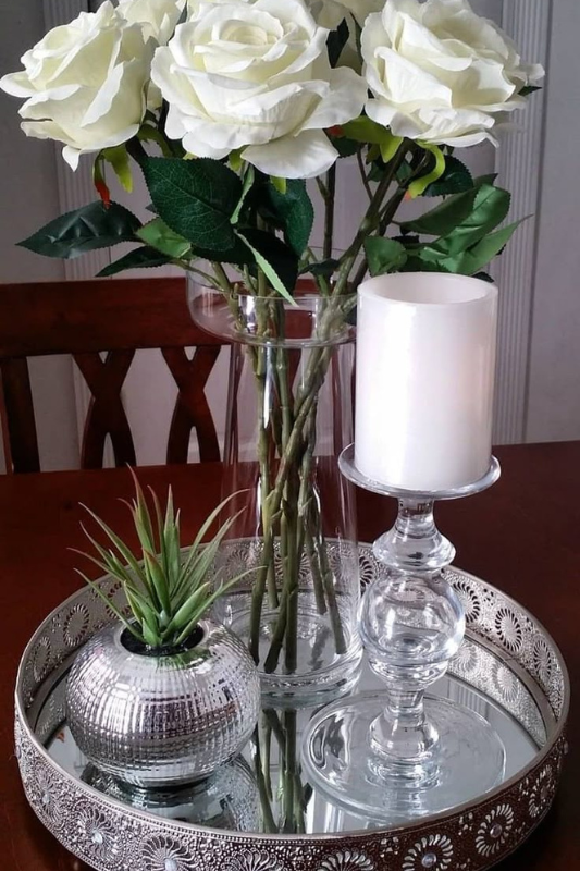 Glamorous centerpiece featuring a silver mirrored tray with a tall glass vase of artificial white roses, a glass candle holder with a white pillar candle, and a metallic vase holding a faux succulent, all styled on a dark wood dining table. 