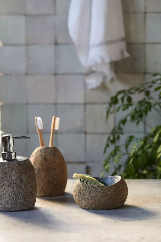 Natural stone bathroom accessories set including a soap dispenser, toothbrush holder, and soap dish, displayed on a neutral countertop with greenery and soft lighting for a serene, spa-like aesthetic. 