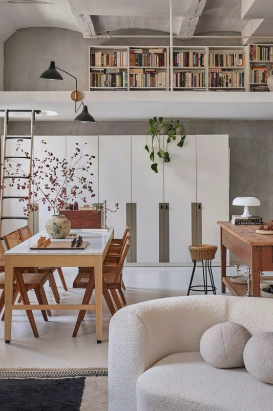 Loft-style studio apartment with overhead bookshelf storage, mid-century dining table, white cabinetry, and cozy boucle sofa for a stylish small space layout








