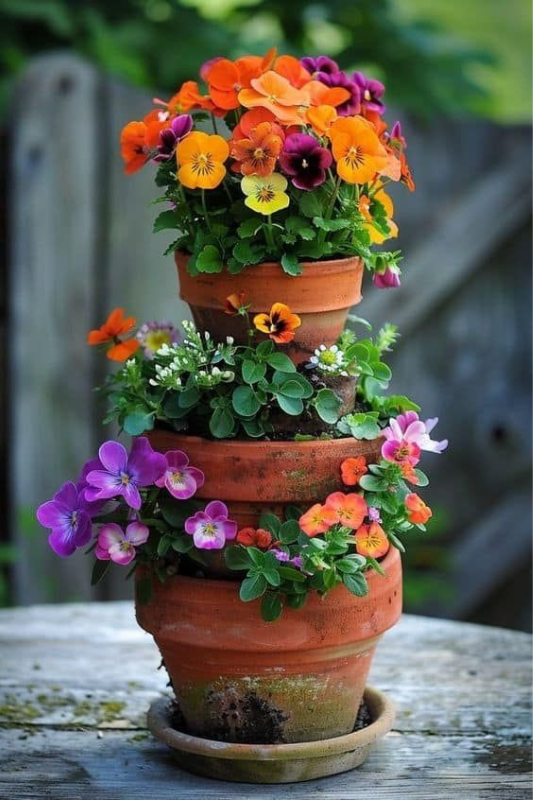 Whimsical tower of aged terracotta pots arranged in a cascading tiered formation, planted with vibrant pansies and assorted trailing flowers, perfect for small garden tabletops. 
