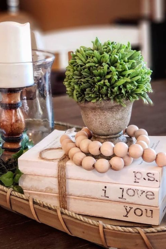 Cozy rustic tray centerpiece featuring a faux potted topiary, stacked decorative books tied with twine and stamped with "P.S. I love you," a wood-beaded garland, and a glass hurricane candle holder with greenery accents. 