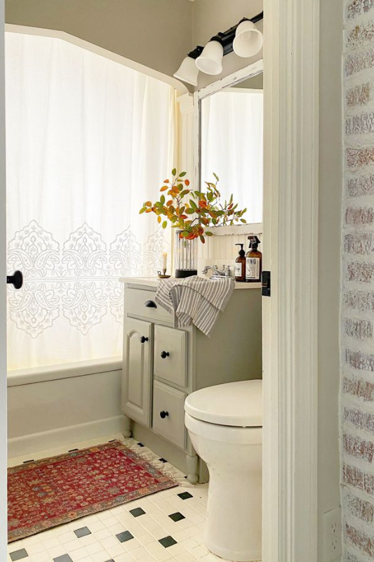 Charming small bathroom featuring a sage green vanity with black hardware, vintage red accent rug, patterned white shower curtain, black-and-white checkered tile floor, and fall-inspired florals styled in a glass vase on the counter. 