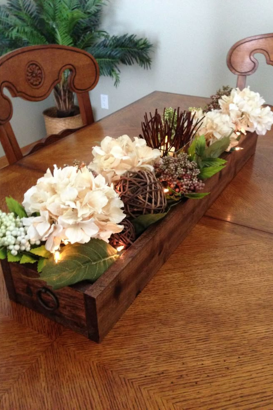 Rustic dining table centerpiece featuring a long wooden box filled with cream hydrangeas, faux berries, twig spheres, green leaves, and warm white fairy lights, styled on a wooden table with carved chairs for a cozy and elegant look. 