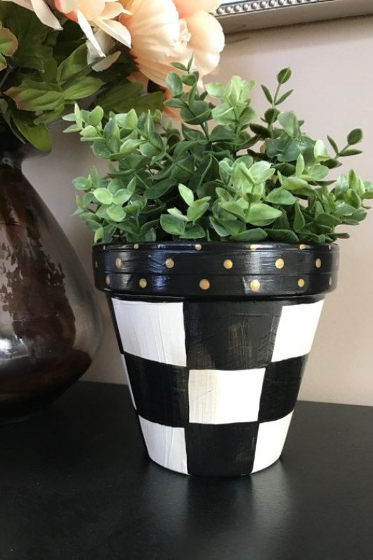 Hand-painted black and white checkered terracotta planter pot with gold polka dot rim, styled with lush faux greenery. This DIY buffalo plaid flower pot adds a charming farmhouse touch and bold visual contrast to modern or rustic home decor displays.