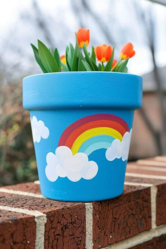 Bright blue hand-painted flower pot featuring a colorful rainbow and white fluffy clouds, filled with blooming orange tulips. Perfect for spring or kids’ garden crafts, this cheerful DIY planter adds a playful pop of color to outdoor patios, windowsills, or garden ledges. Ideal for whimsical and creative decor themes.