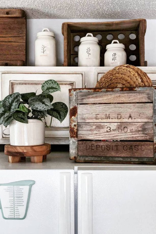 A cozy farmhouse-style kitchen scene featuring the top of a white refrigerator decorated with a white potted plant on a wooden stand, a rustic wooden crate labeled "E.M.D.A. DEPOSIT CASE," woven tray, and vintage white canisters with botanical labels displayed in wooden crates above distressed white cabinets.
