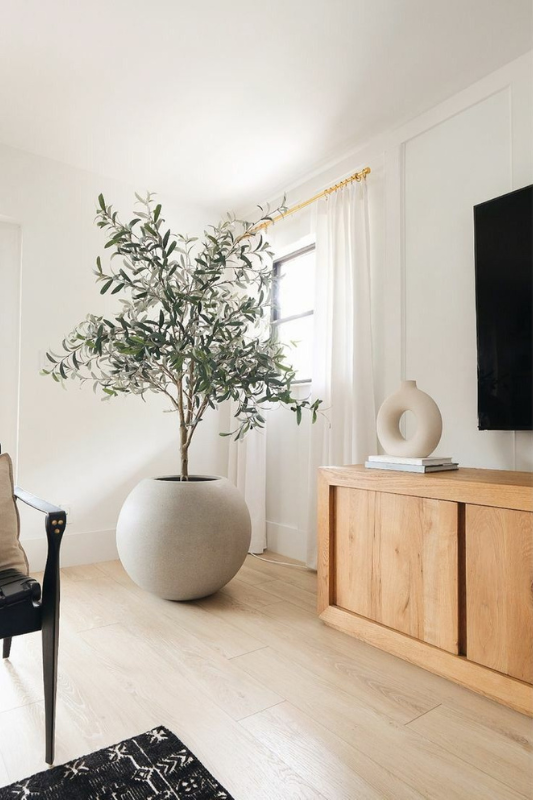 Minimalist modern living room featuring a large potted faux olive tree in a round stone planter, light wood floors, white walls with paneling, a wooden sideboard, and gold curtain hardware. This serene neutral interior is styled for an airy, natural aesthetic that blends Scandinavian and Japandi design elements.