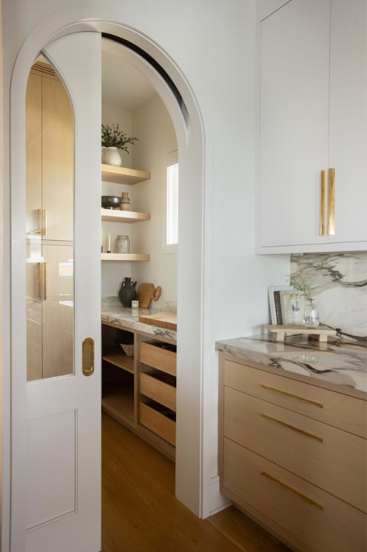 Elegant arched pocket door revealing a pantry with wooden shelves and marble countertops in a modern kitchen.