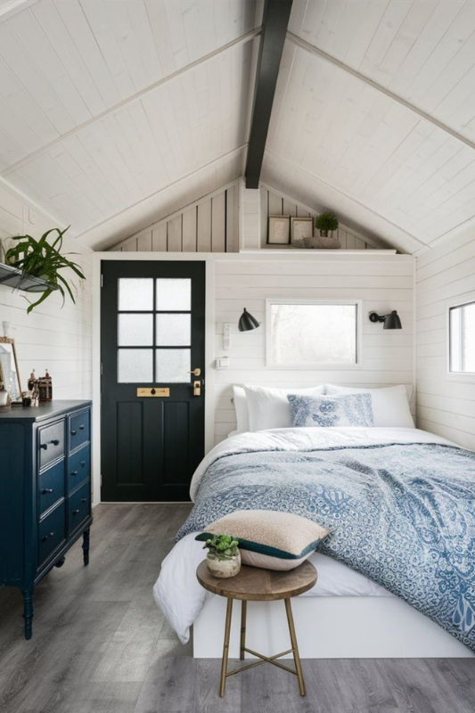 Stylish white-paneled she shed bedroom with vaulted ceiling, black front door, blue vintage dresser, and bed with blue-and-white patterned bedding.